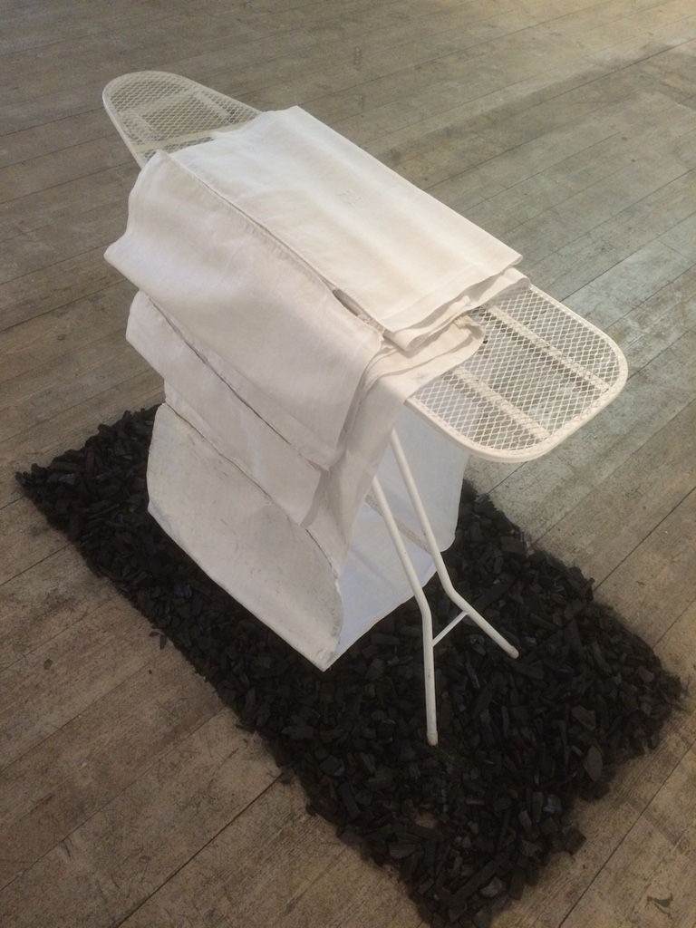 Strykbräda med vita handdukar står på en rektangulär yta av kolsot. Verk av Christina Göthesson 